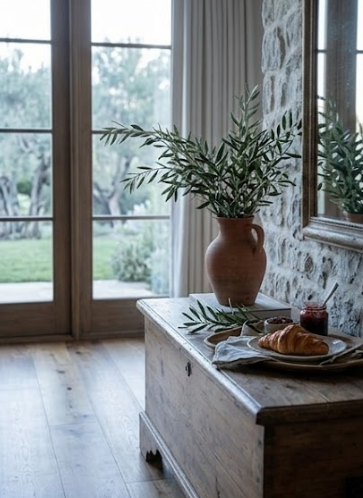 Plan rapproché sur une commode en bois patiné dans une chambre d'hôtes en pierre. Sur un plateau, un croissant doré, de la confiture et un vase en terre cuite avec une branche d'olivier devant une grande fenêtre.